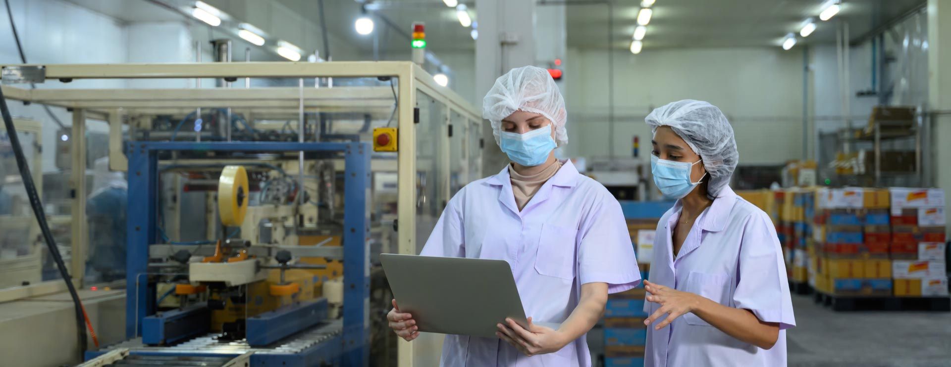 Zwei Frauen in Schutzkleidung, Maske und Haarnetz schauen auf ein Tablet, während sie in einer Produktionshalle stehen.