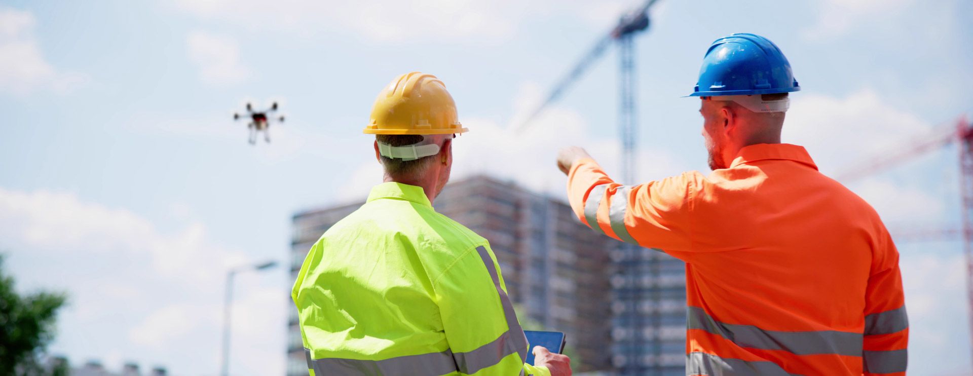 Zwei Männer in Warnjacken und Sicherheitshelmen stehen auf einer Baustelle. Einer von ihnen zeigt auf eine Drohne