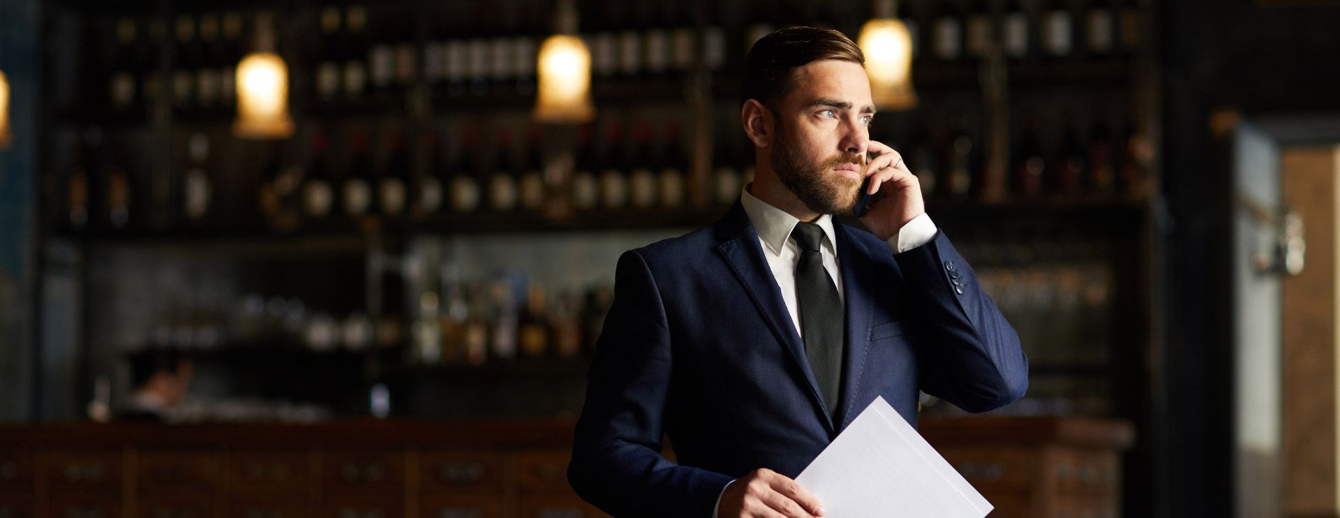 Ein Mann im Anzug steht in einem Restaurant und hält ein Klemmbrett während er sich ans Ohr fasst