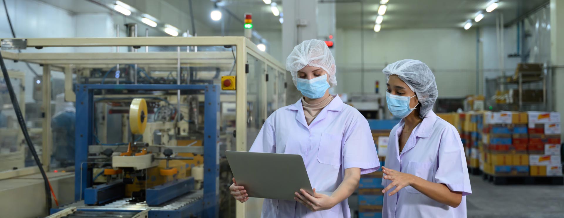 Zwei Frauen in Schutzkleidung, Maske und Haarnetz schauen auf ein Tablet, während sie in einer Produktionshalle stehen.