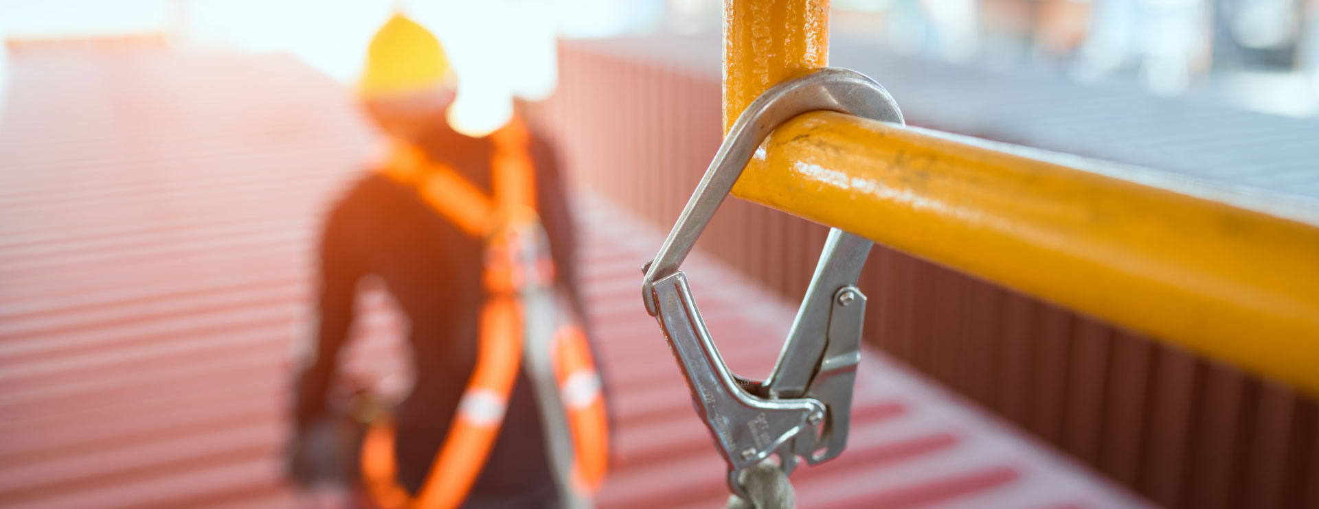 Ein Karabinerhaken im Vordergrund schützt eine Person, die auf einem roten Dach arbeitet.