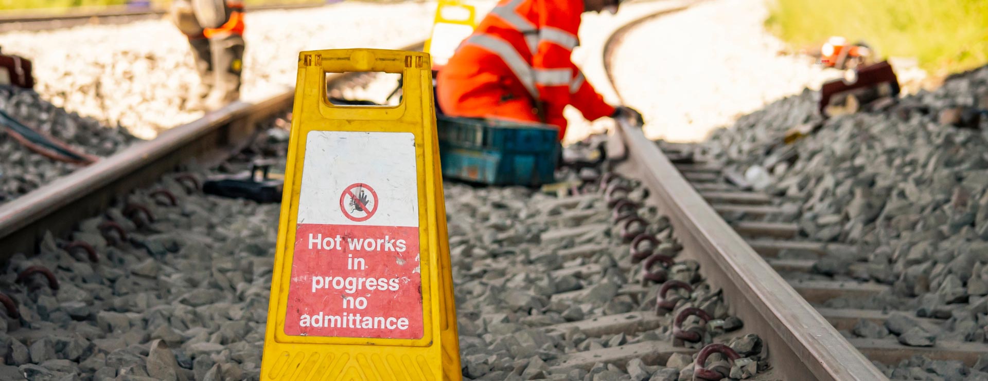 Vor Bauarbeitern, die an einem Gleis arbeiten steht ein gelbes Warnschild.