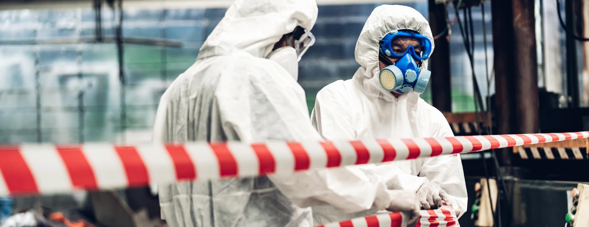 Zwei in Schutzkleidung gehüllte Menschen mit speziellen Atemmasken sperren einen Bereich ab.