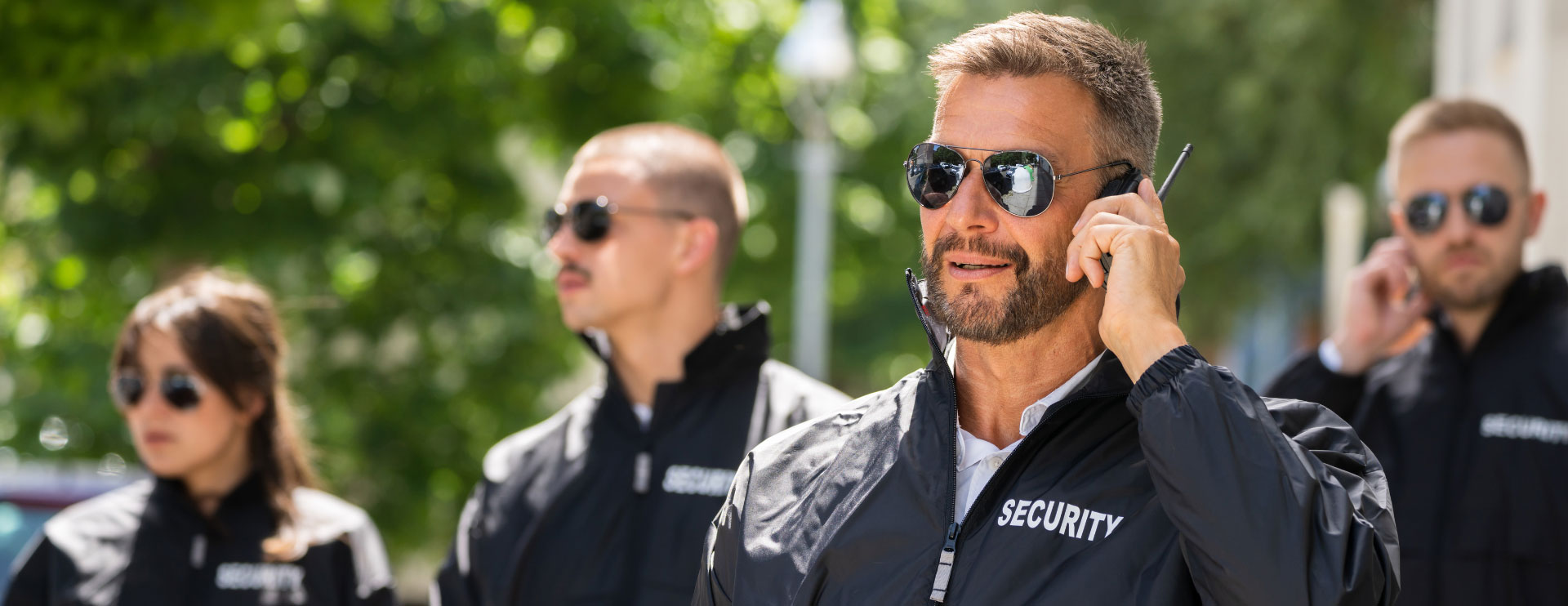 Vier Personen in Security-Kleidung bewachen eine Grünanlage. Alle haben Sonnenbrillen auf und zwei halten je ein Walkie-Talkie ans Ohr.